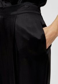 Un pantalon en satin noir met en valeur une texture lisse avec une taille ajustée et des poches latérales, souligné par des plis subtils pour une touche de détail supplémentaire.