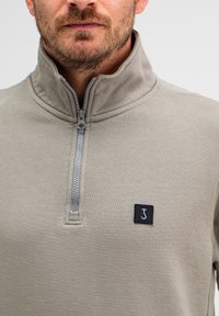 Hellgrauer Pullover mit einem Halb-Reißverschluss-Kragen und einem schwarzen quadratischen Logo-Patch. Der Stoff scheint weich mit einer subtilen Textur zu sein.