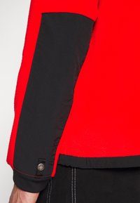 Czerwony polarowy płaszcz z czarną tkaniną na dolnej części rękawa i u dołu, z detalem srebrnego zatrzasku w okolicy nadgarstka, noszony z czarnymi spodniami.