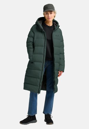 Person wearing a long dark green puffer jacket, black cap, black shirt, blue jeans, and black shoes standing against a plain background.