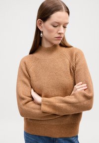 Pulover tricotat, cu textură ribbed, de culoare maro scorțișoară, cu guler înalt și mâneci raglan. Textură moale cu talie ajustată. Stilizat cu blugi albaștri.