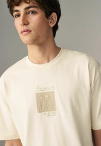 Cremfarbnes Baumwoll-T-Shirt mit einem kleinen, gestickten, von Picasso inspirierten Gesichtsmuster in Beige. Mit Rundhalsausschnitt und kurzen Ärmeln. Schlichte, klare Form.