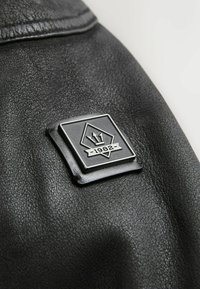 Gros plan sur une veste en cuir noir avec un patch en métal de forme carrée, comportant un motif en forme de diamant, une couronne et le texte "1982". La texture est granuleuse.