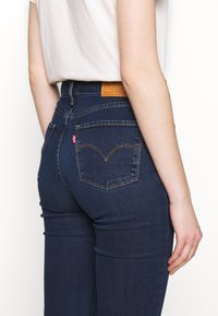 Jeans de mezclilla azul oscuro con una cintura alta, bolsillos traseros con costuras distintivas y una etiqueta de cuero marrón en la cintura.