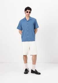 Μπλε κοντομάνικο πουκάμισο με κουμπιά από βαμβάκι, συνδυασμένο με άσπρα σορτς. Μοντέλο που φορά μαύρα παπούτσια και γυαλιά ηλίου, στέκεται μπροστά σε λευκό φόντο.