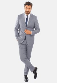 Abito grigio chiaro con un design aderente, giacca monopetto, due tasche frontali e pantaloni abbinati; abbinato a una cravatta scura e scarpe nere lucide.