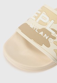 Sandale en caoutchouc beige avec une bride texturée portant le texte en relief "MILANO" et un semelle lisse. Le design inclut un motif camouflage.