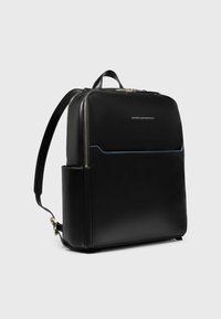 Mochila de cuero negro con una forma rectangular delgada, que presenta un bolsillo frontal con costuras en azul y detalles de cremalleras doradas.