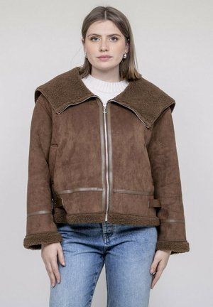 Jeune femme portant une veste en peau lainée marron avec un grand col et un jean bleu, debout devant un fond clair uni.