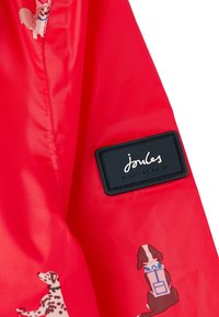 Röd vattentät jacka med en slät textur, prydd med hundtryck och en svart märkestagg som läser "Joules Right as Rain."