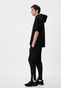 Schwarzes Kapuzenshirt mit lockerer Passform, kombiniert mit schwarzen taillierten Cargo-Hosen mit Reißverschlüssen an den Bündchen. Getragen mit schwarzen Sneakers.