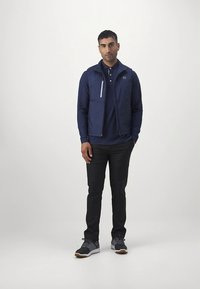 Granatowa kurtka z wysokim kołnierzem i zamkiem błyskawicznym, w zestawieniu z granatową koszulką polo, czarnymi spodniami i szarymi butami sportowymi. Prosty design, bez wzorów.