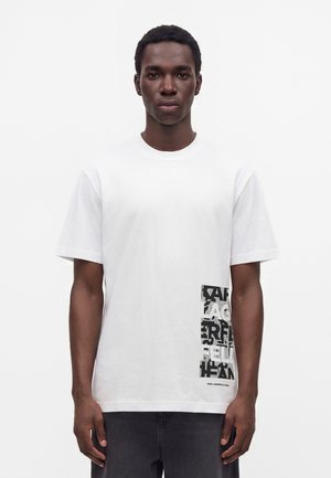 Νέος άνδρας με σκουρόχρωμο δέρμα που φοράει λευκό μπλουζάκι με μαυρόασπρο αφηρημένο μπλοκ κειμένου στο κάτω δεξί μέρος.