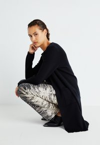 Svart stickad cardigan med långa ärmar, kombinerad med tigerprickiga vida byxor i beige och svart, kompletterad med svarta spetsiga skor.