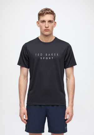 Homme debout portant un t-shirt noir Ted Baker Sport et un short bleu marine sur un fond blanc uni.