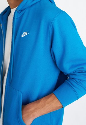 Personne portant un sweat à capuche zippé Nike bleu vif, main dans la poche avant, t-shirt blanc en dessous, gros plan sur le haut du torse et le bras.