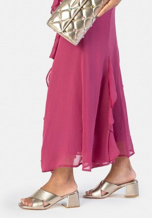 Mujer con un vestido magenta fluido que sostiene un clutch dorado acolchado, llevando sandalias doradas de tacón con tiras cruzadas.