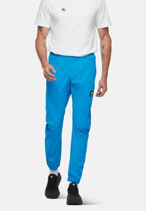 Mand iført lyseblå joggingbukser med en logo-patch på låret, en hvid t-shirt, hvide sokker og sorte sneakers.