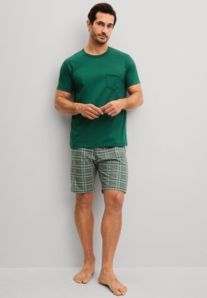 Man standing barefoot wearing a green short-sleeve shirt with a chest pocket and green plaid shorts against a gray background.