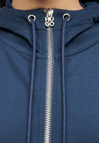 Μπλε σκούρο φούτερ με ασημένιο φερμουάρ, που διαθέτει έμβλημα στην τσόχινη ταινία και κορδόνια. Ομαλό ύφασμα με απαλή υφή και απλή σχεδίαση.