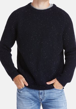 Personne portant un pull en maille bleu foncé moucheté et un jean bleu clair avec les mains dans les poches, cadrée au niveau du cou et des cuisses.