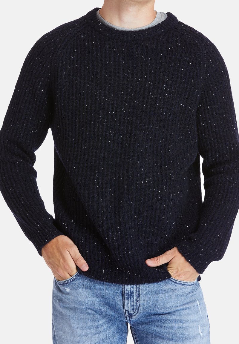 Personne portant un pull en maille bleu foncé moucheté et un jean bleu clair avec les mains dans les poches, cadrée au niveau du cou et des cuisses.