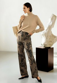 Vrouw in een beige coltrui, met een patroon op brede broekspijpen, zwarte puntige schoenen, die een beige tas vasthoudt en bij een abstracte witte sculptuur staat.