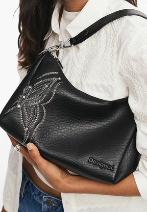Sac à bandoulière en cuir noir avec motif papillon brodé blanc, clous argentés et logo "Desigual", porté avec un chemisier blanc texturé.