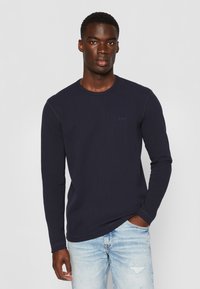 Marineblaues Langarm-T-Shirt aus strukturiertem Stoff, mit Rundhalsausschnitt und dezentem Logodetail auf der Brust, kombiniert mit hellen, stark strapazierten Jeans.