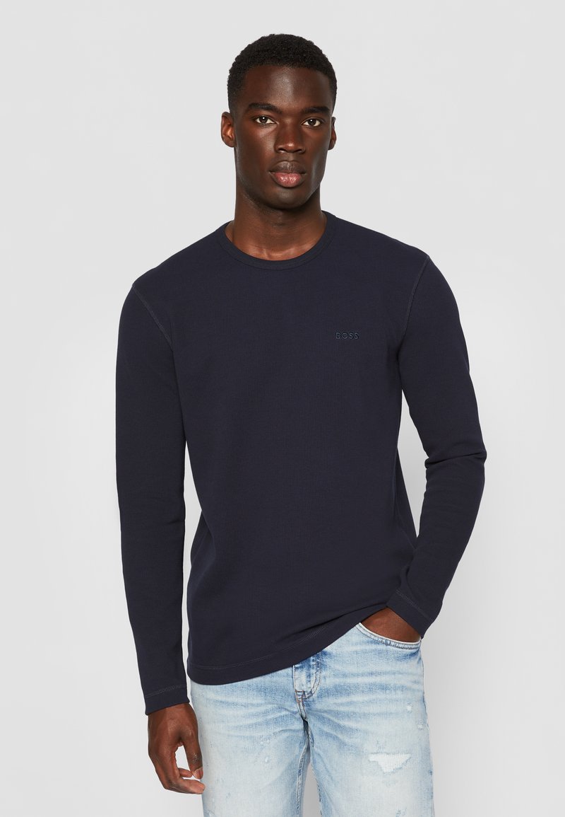 Marineblaues Langarm-T-Shirt aus strukturiertem Stoff, mit Rundhalsausschnitt und dezentem Logodetail auf der Brust, kombiniert mit hellen, stark strapazierten Jeans.