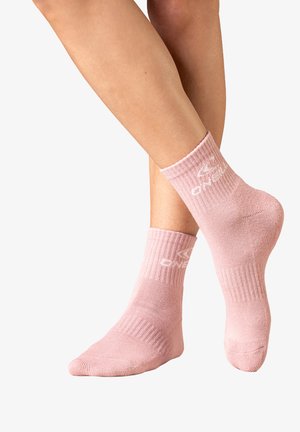 Ben gekruist met lichtroze geribbelde enkelsokken met wit merkenlogo op de zijkanten tegen een witte achtergrond.
