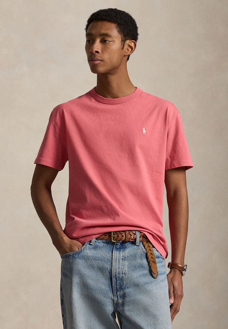 Νέος άνδρας που φοράει ροζ κοραλλί κοντομάνικο πουκάμισο, ανοιχτό μπλε τζιν με καφέ πλεγμένη ζώνη και καφέ ρολόι χειρός.