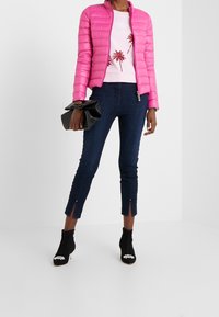 Chaqueta acolchada rosa, sudadera blanca con gráfico de palmeras, jeans de mezclilla oscuros recortados con aberturas laterales y botines negros con adornos.
