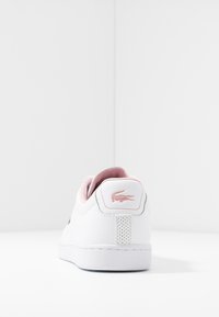 Sneaker bianca con un accento rosa sul tallone posteriore, dotata di un logo e dettagli testurizzati, su uno sfondo bianco uniforme.