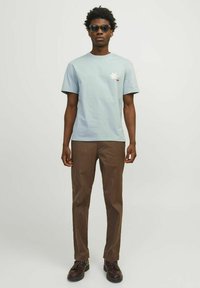Camiseta de algodón azul claro con mangas cortas, que presenta un pequeño diseño bordado; combinada con pantalones marrones y zapatos oscuros.