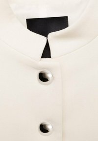 Vêtement couleur crème avec un col rond, doté d'une étiquette en tissu noir et de deux boutons-pression métalliques. Texture lisse.