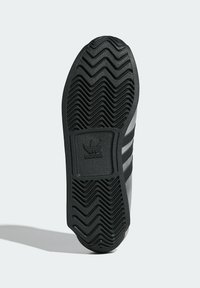 Black rubber shoe sole with a zigzag tread pattern, featuring three black stripes and an embossed logo section.