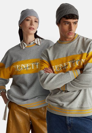 Due modelle che indossano felpe Benetton a righe grigie e gialle e berretti lavorati a maglia grigi, in piedi fianco a fianco su uno sfondo bianco.