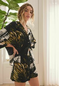 Conjunto de blusa de manga corta con botones y pantalones cortos con estampado tropical en negro, blanco y verde; tela suave con un corte relajado y mangas anchas.