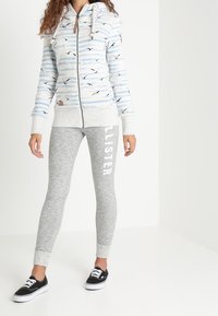 Sudadera con cremallera a rayas azul claro y blanca con patrones de pájaros, combinada con leggings gris jaspeado que llevan texto blanco, y zapatillas negras.