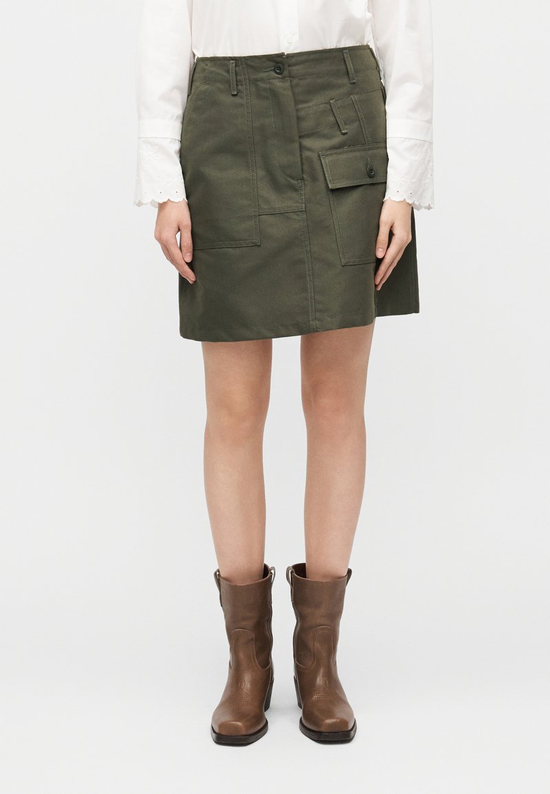 Πράσινη φούστα από βαμβάκι με ορατές ραφές, πολλαπλές τσέπες και εφαρμοστή μέση. Συνδυάζεται με καφέ μποτάκια αστραγάλου.