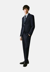 Mornarski odijelo s prilagođenim dizajnom, jednorednim sakoom, reverima urezanim u obliku slova "V" i odgovarajućim hlačama. Uključuje svijetloplavu košulju i prugastu kravatu.