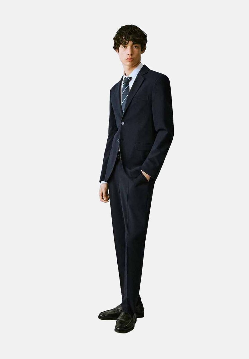 Mornarski odijelo s prilagođenim dizajnom, jednorednim sakoom, reverima urezanim u obliku slova "V" i odgovarajućim hlačama. Uključuje svijetloplavu košulju i prugastu kravatu.