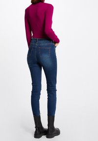Femme portant un jean slim bleu foncé, des bottines noires et un haut à manches longues magenta, debout avec une main dans la poche sur un fond blanc.