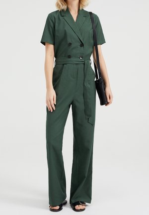 Grüner Jumpsuit aus Baumwollmischung mit kurzen Ärmeln, zweireihiger Vorderseite, weitem Bein und passendem Bindegürtel. Schwarze Umhängetasche und Sandalen.