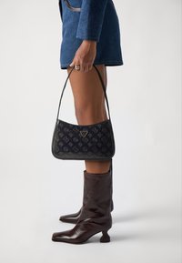 Sort læderhåndtaske med præget logo og blomstermønstre, der har en buet form og en tynd skulderrem, præsenteret sammen med brune læderknæhøje støvler.