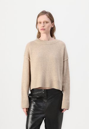 Beige kort genser laget av mykt strikket materiale, med en bred rund hals og ribbestrikkede mansjetter, kombinert med svarte glinsende lærbukser.