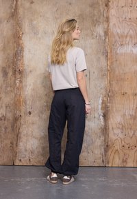 Kobieta o długich blond włosach stoi twarzą do drewnianej ściany, ubrana w beżowy t-shirt, czarne luźne spodnie i brązowe trampki z białymi paskami.