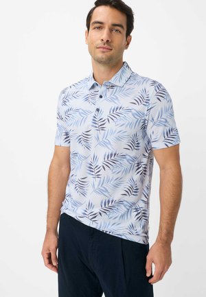 Mann trägt ein kurzärmeliges, hellblaues Poloshirt mit dunkelblauem Blattmuster und dunkle Hose, steht vor weißem Hintergrund.