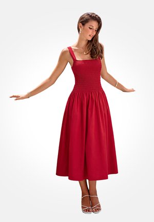 Kobieta w czerwonej sukience midi bez rękawów z marszczonym gorsetem i plisowanym dołem, ubrana w białe sandały na obcasie z paskami, stojąca z lekko uniesionymi ramionami.